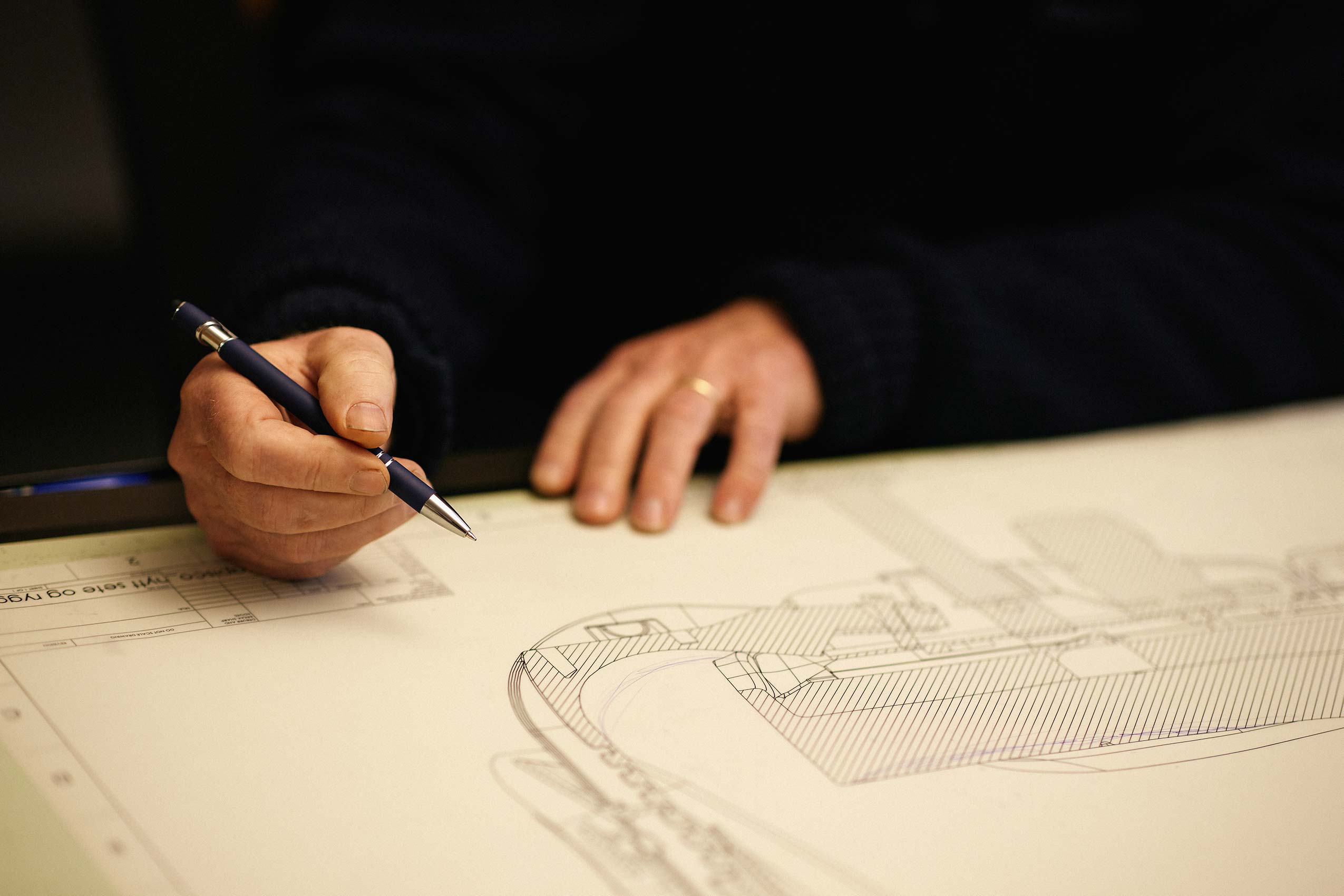 a close up of two hands, one holding a pen, the other ontop of a large piece of paper with a sketch of the inner workings of a chair design. the person has a ring on their finger, and is wearing all black. the pen is black