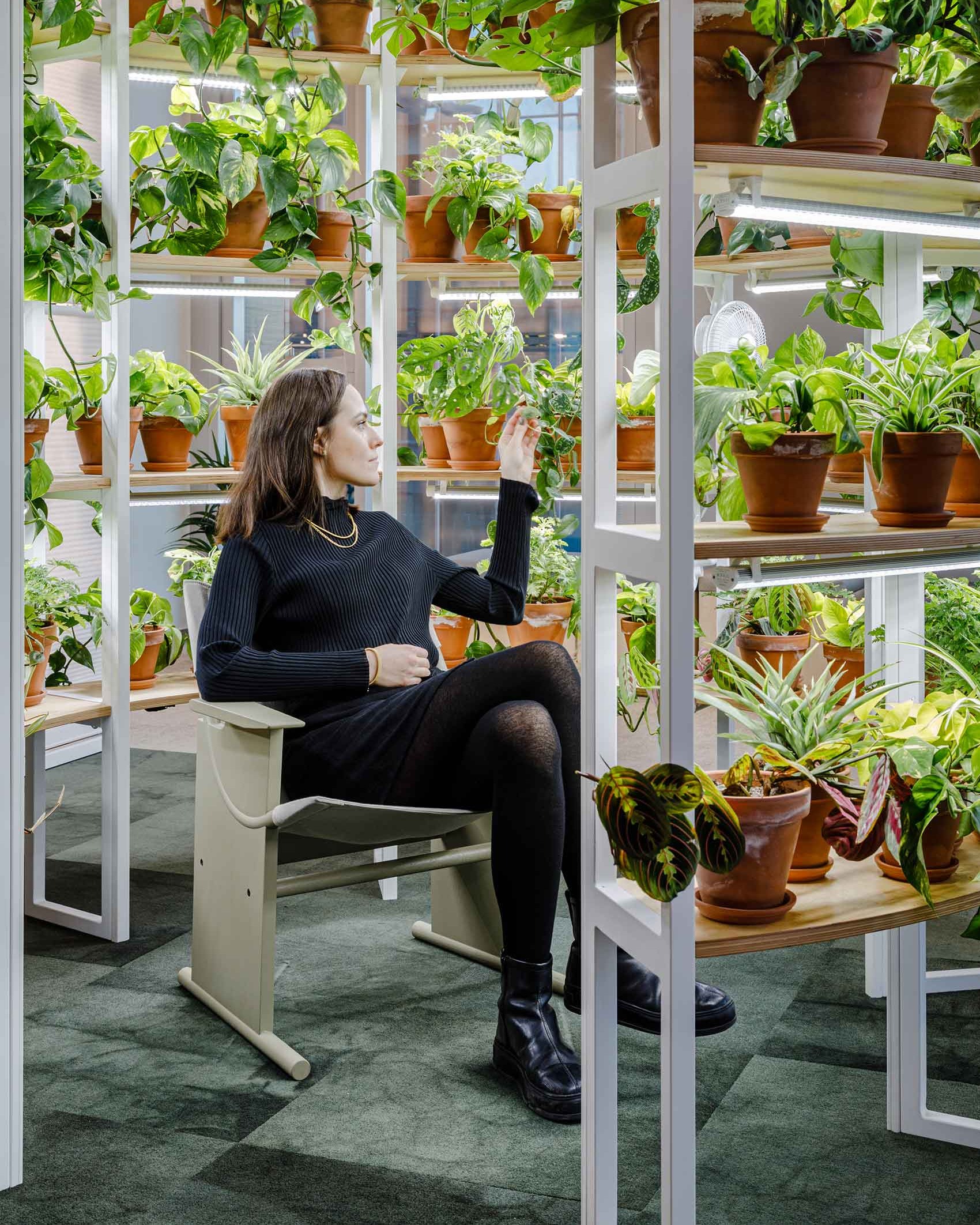 A lady sat in a chair surrounded by green plants in pots on a circle of shelving units