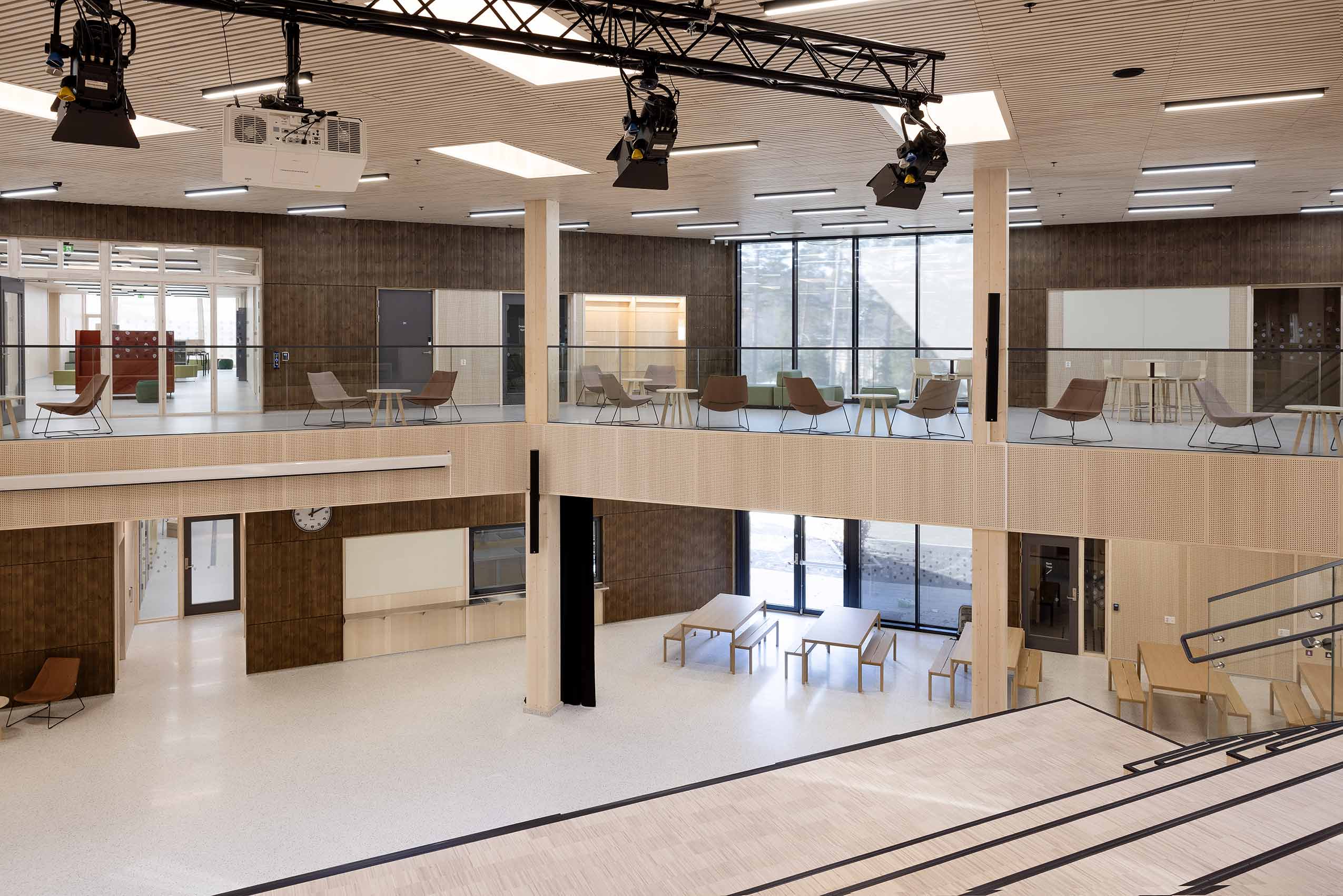 Central social space at school, with tables and bences on the ground floor and Profim Chic lounge chairs on the second floor, visible on a walkway around the top. the floor and walls are wooden and bright, with ceiling lights visible and a projector 
