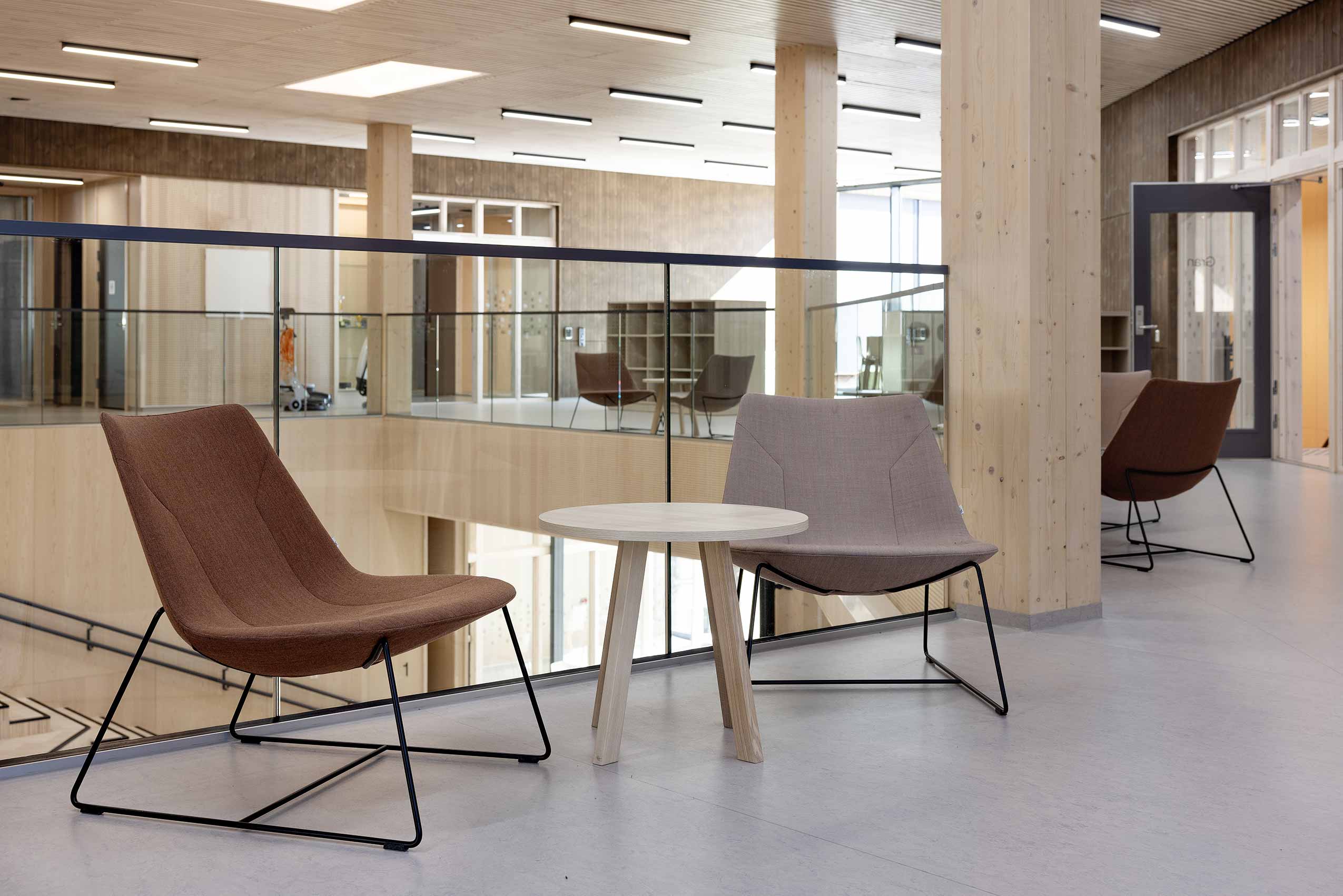 Two Profim Chic chairs facing the camera with a small table between them, on a second floor walkway at a school, with a glass panel overlooking the first floor, and wooden clad walls and beams