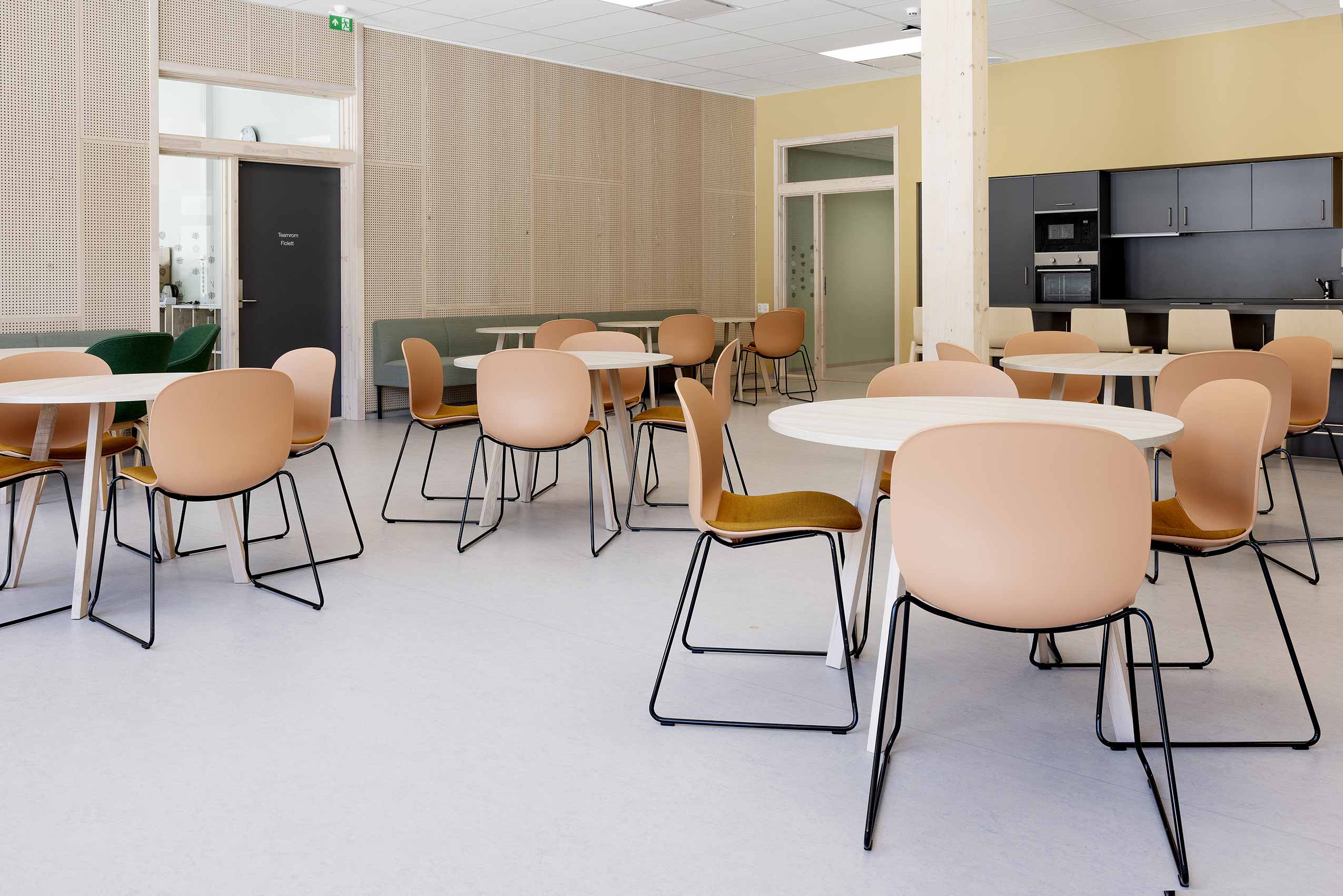 Interior of HOV school, with round tables spaced out with pink Profim noor chars in groups of 4 around each table, the walls are wooden and the room is sparcely filled