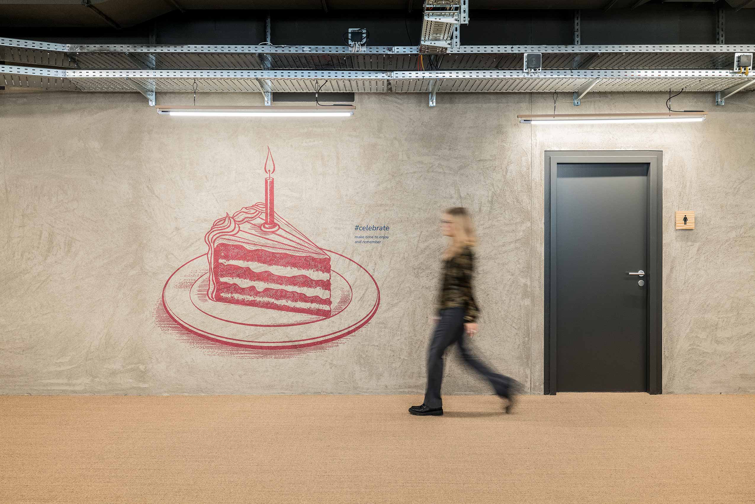 hallway at I-City, with a red birthday cake slice artwork on a concrete wall, a door and a lady walking past
