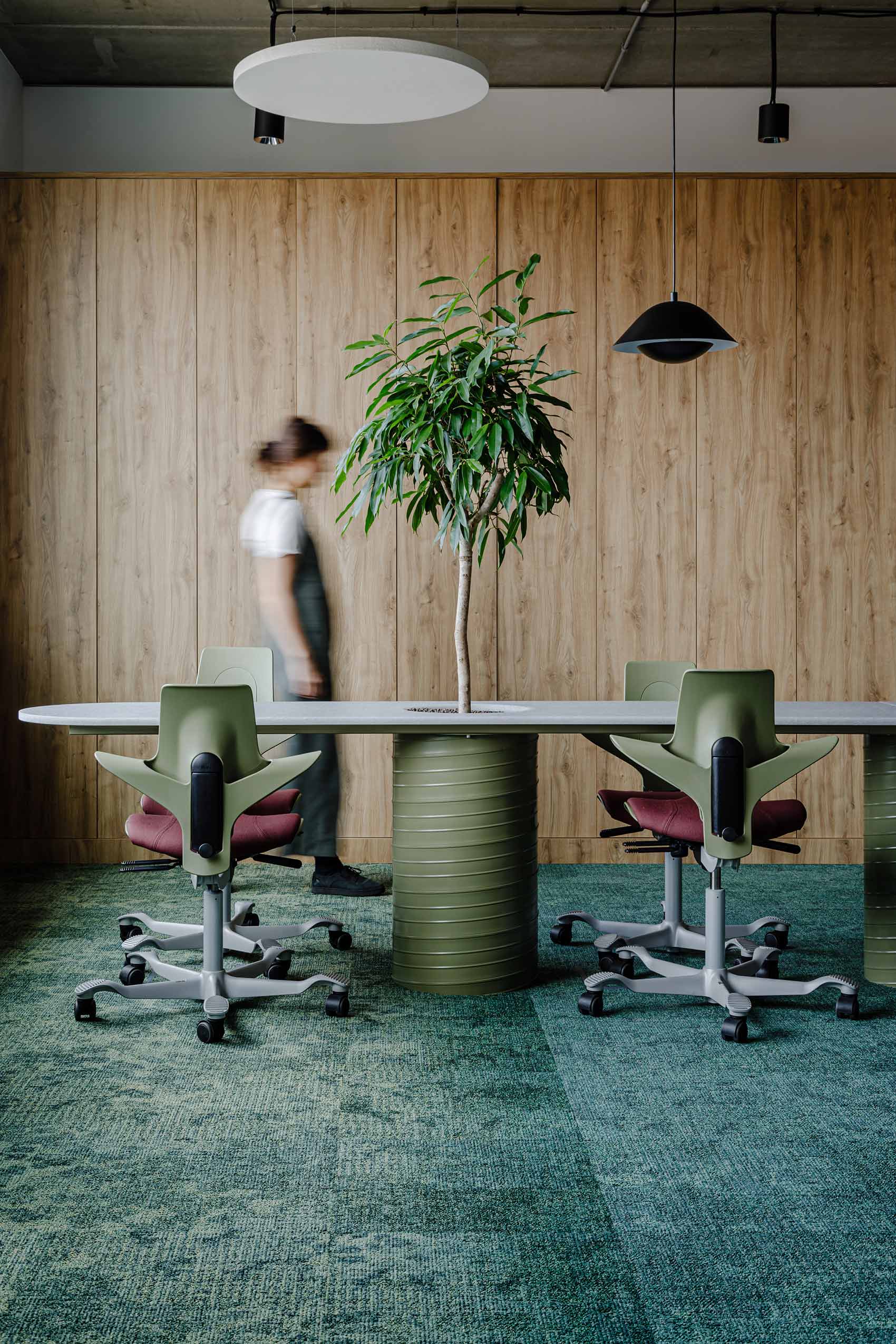 artful shot of an office with green HÅG Capisco Puls chairs a table with a plant on it and a lady standing hbehind it. the wall is wooden panelled