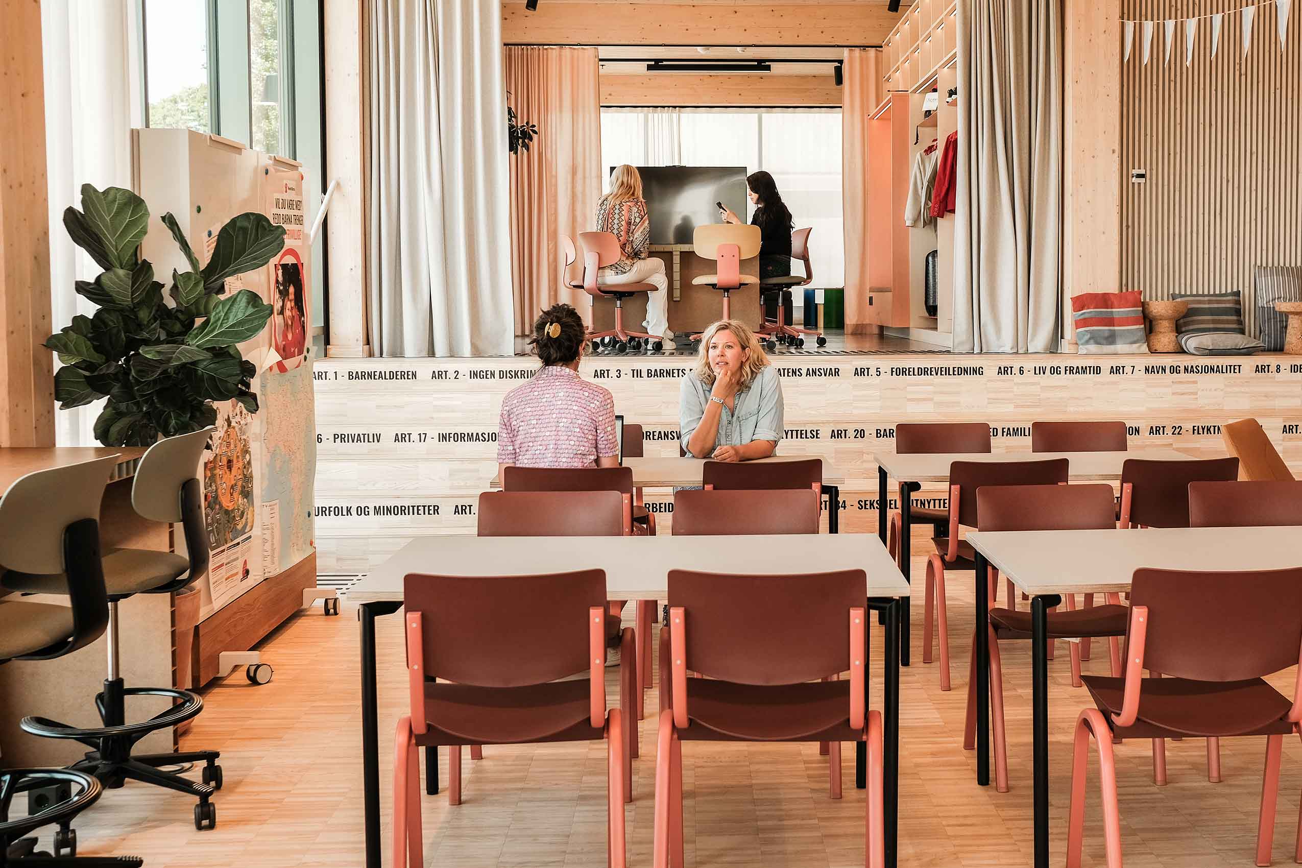 Angle 2 of two people talking sat no brick red HÅG celi chairs at a table, in Redd Barna's Oslo office