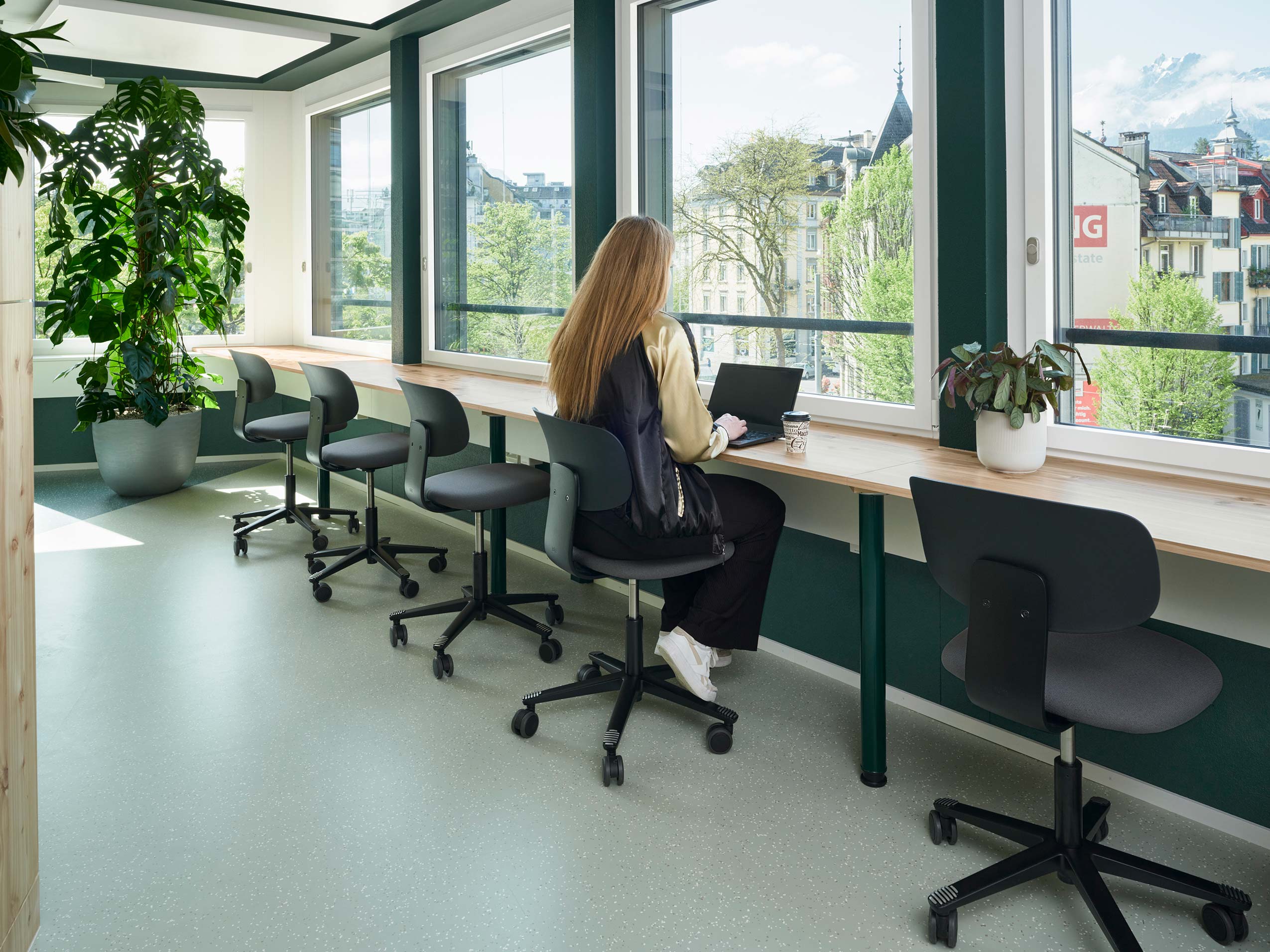 a bright room with big windows along the wall. inside is a high simple table attahced to the wall with a row of 5 HÅG Tion chairs lined up along the wall. a lady is sat working on a laptop on the middle chair