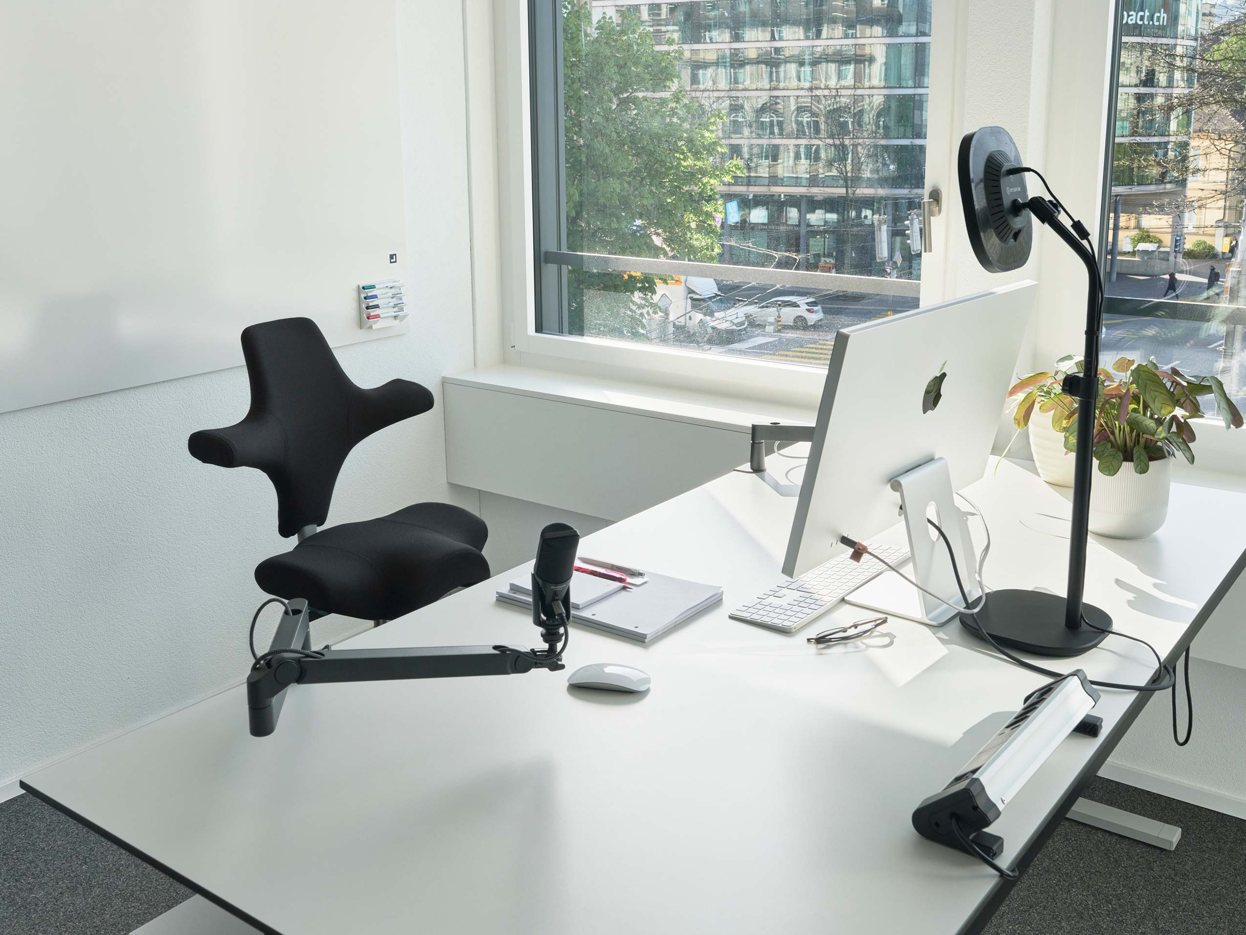 HÅG Capisco chair in black, behind a white desk with an apple mac screen and a window in the background