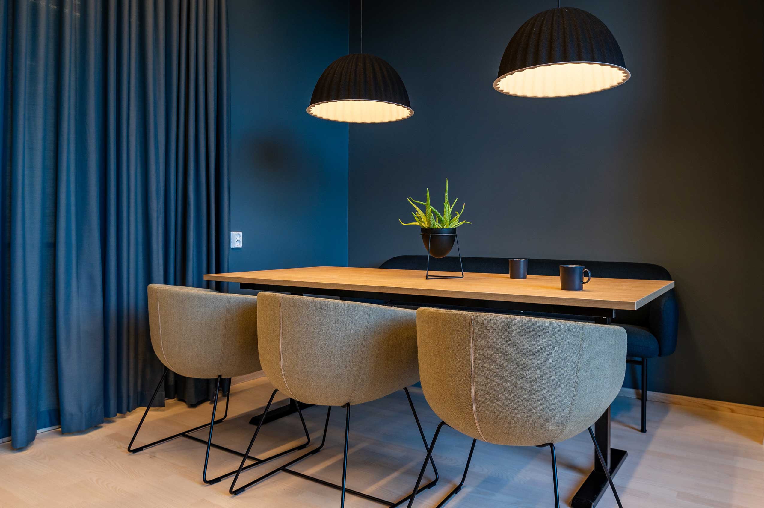 3 Profim Nu chairs facing a blue wall with a long rectangular table infront of them. there is a blue curtain on the left of the frame and 2 pendant lights hanging above the table